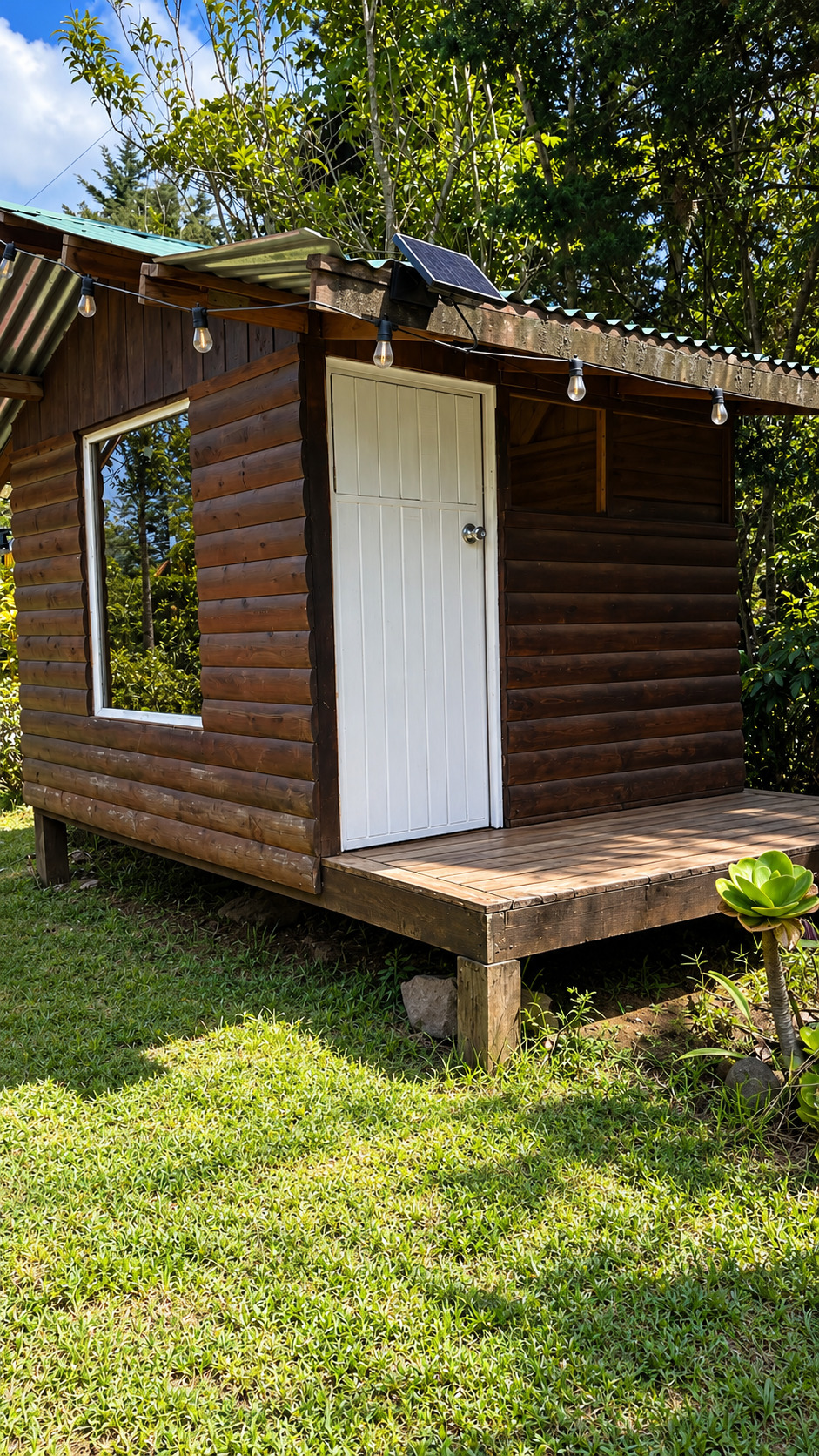 Casita de troncos con puerta blanca, luces colgantes y panel solar en el jardín